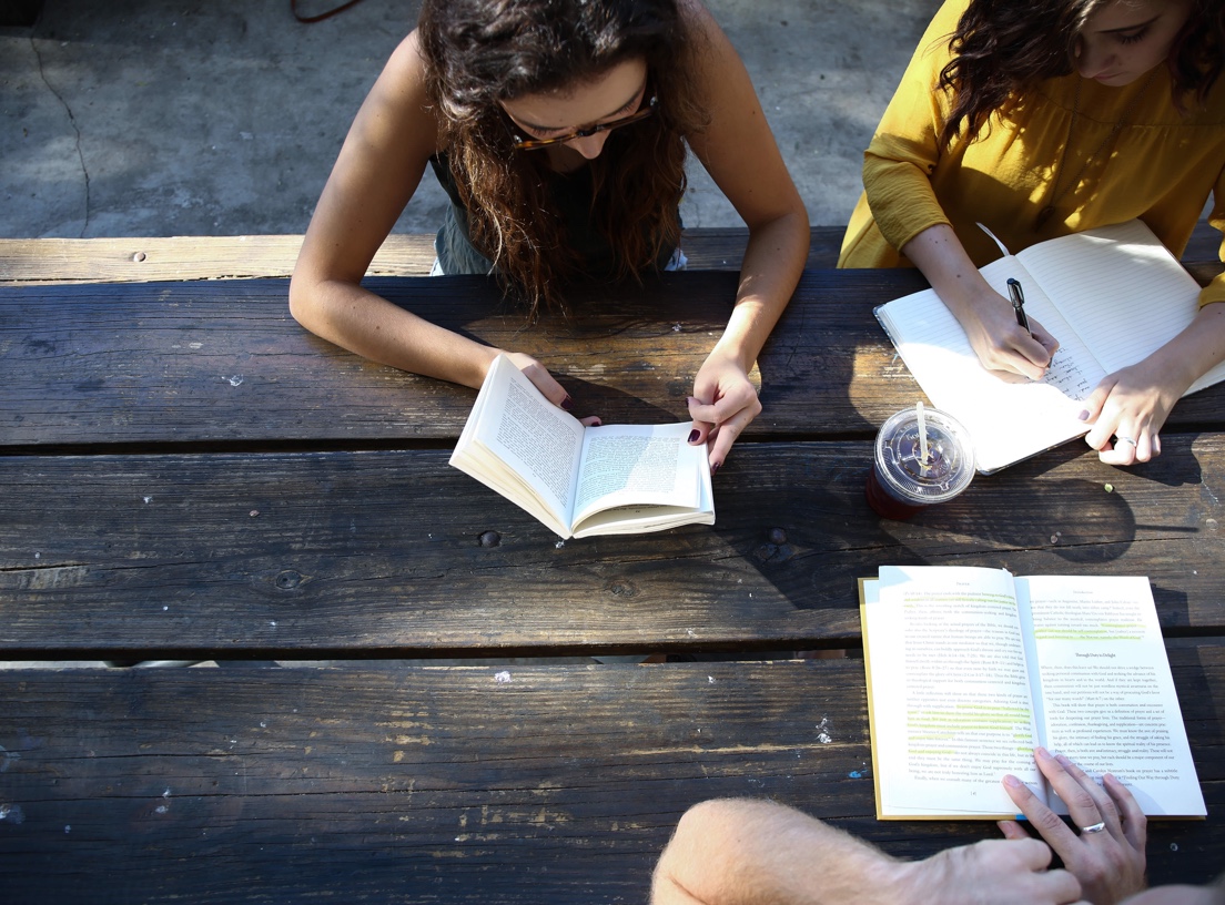 Three college students at a picnic table, reading from books and taking notes. A student has a drink in a clear, plastic cup with a lid and straw.